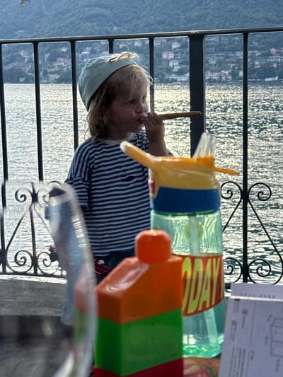 A young child wearing a blue baseball cap and a blue and white striped t-shirt is holding a brown object to their mouth, seemingly pretending to smoke. They are standing near a black metal railing, behind which is a body of water reflecting sunlight, with a town visible on the distant hillside. In the foreground, out of focus, are colorful building blocks and a water bottle. The scene appears to be outdoors, possibly on a balcony or patio overlooking a lake, with the town of Pognana Lario, Italy, in the background.