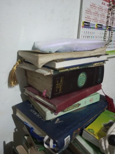 A stack of religious books, including copies of the Quran, is seen in the image. One book has "YAASIIN HAUHILAN" printed on its spine. The books vary in color, with blue, red, and dark brown covers visible. A gold tassel hangs from one of the books. A calendar is partially visible in the background, showing September and October. The word "JUAL" is partially visible on the calendar. A string of beads is hanging from the calendar.
