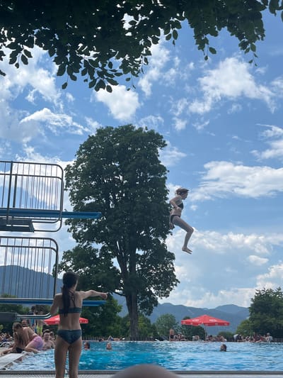 In Oberammergau, Germany, a bright, cloud-filled sky forms the backdrop for a refreshing summer scene at an outdoor swimming pool. The clear blue water of the pool is dotted with swimmers and sunbathers relaxing on the poolside. On the right side of the frame, a young woman in a black bikini is captured mid-air, mid-dive from a high diving board, her body contorted as she prepares to enter the water. To the left, another young woman, also in a bikini, stands at the edge of the pool, pointing towards the diving woman. In the background, a lush green forest with a towering tree dominates the landscape, with rolling hills and mountains visible in the distance. Red umbrellas, providing shade for swimmers, are scattered around the pool area.