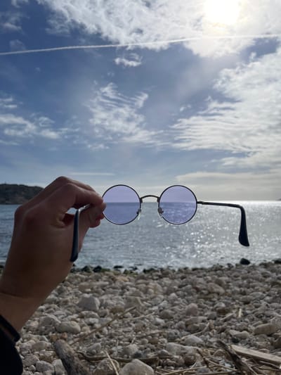 The image captures a detailed view of a coastal scene in L'estartit, Spain. In the foreground, a human left hand grasps a pair of round-framed sunglasses featuring dark metal frames and purple-tinted lenses. The hand is visible from the wrist, which is adorned with a dark cuff. The right temple arm of the sunglasses extends horizontally. The immediate environment beneath the sunglasses consists of a pebble beach, characterized by light grey and tan stones, with small pieces of driftwood scattered across the surface. Behind the sunglasses, the calm Mediterranean Sea is visible, reflecting abundant sunlight as shimmering points on its surface. To the far left, a blurred, dark landmass, consistent with a coastal hill or cliff, emerges from the water. The upper sky displays a mix of blue and scattered white clouds, including a prominent white aircraft contrail stretching horizontally across the upper left. A bright, partially obscured sun is located in the upper right portion of the sky, serving as the primary light source.