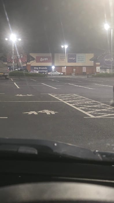 The image depicts a nighttime scene of a retail park parking lot in Newry, United Kingdom, viewed from inside a vehicle, with the dark dashboard partially visible in the foreground.
The midground features a dark asphalt parking lot, largely empty of vehicles. White painted lines delineate individual parking spaces. Multiple white stenciled symbols for "parent and child" parking, depicting two stylized human figures, are visible on the asphalt. A rectangular area marked with white diagonal hatching is also present.
In the background, commercial buildings are brightly illuminated by multiple overhead streetlights, causing lens flare. The building on the left displays a "B&M" logo and the text "Big Brands Big Savings," with a red and white striped motif above. Adjacent to it, another building presents the "Currys" logo, alongside text including "Enjoy support hub." and "talk tech. with tech people." This structure features a lower facade of reddish-brown brick. A silver sedan is visible in the distance, positioned near the retail units. No individuals are discernible.