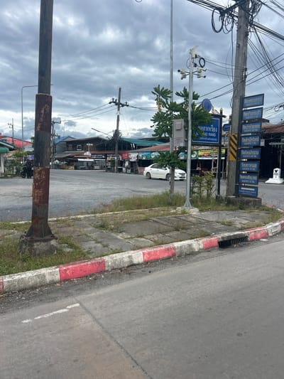 The image shows a street scene in Lan Saka, Thailand. A light-colored sedan is parked on the right side of the road. Several utility poles with numerous wires are visible, along with a traffic light. A signpost with multiple blue signs, some with Thai writing, stands on the right. There is a red and white curb on the left side of the road, leading to a sidewalk with patches of grass. The sky is overcast with gray clouds.
