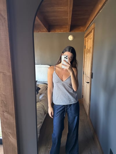 A young adult female is depicted in a full-length mirror selfie within a room in Otego, United States. She stands facing the mirror, holding a light-colored smartphone with both hands to take the photograph, obscuring her face. She wears a light-colored, small-checked camisole with thin straps, dark blue wide-leg pants with a drawstring waist, and a delicate necklace with a small pendant. The reflected room features dark gray walls, a light wood-toned floor, and a wooden beamed ceiling. A light-colored wooden door with a dark handle is visible on the right side of the reflection. To the left, a bed with a white textured headboard and light bedding is visible, with a dark object on its surface. A white circular smoke detector or fixture is mounted on the dark gray wall above the female's head. The mirror has a dark frame with an arched top-left corner, and a prominent wooden beam is visible immediately to the left of the mirror.
