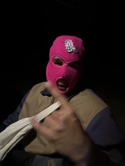 A person wearing a bright pink ski mask with eye holes and a cartoonish logo is depicted. They are holding a white cloth bag. The person's clothing appears to be a jacket or coat with tan and dark blue panels. The background is dark and indistinct. The location of the photo is not discernible from the image and there is no information to indicate this image was taken in Étréchy, France.
