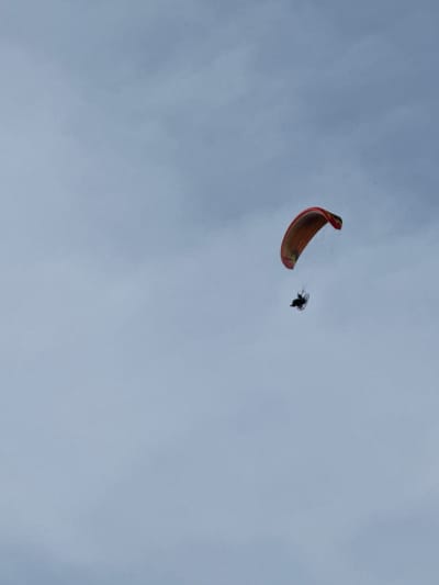 A paramotorist flies in a pale blue, overcast sky. The paramotor wing is primarily orange with red accents and a dark border. The pilot is visible below the wing, seated in a harness with a dark frame and propeller. The scene is devoid of any discernible city features or landscape, suggesting the photograph was taken at a considerable altitude. The location is specified as Žemieji Rusokai, Lithuania.