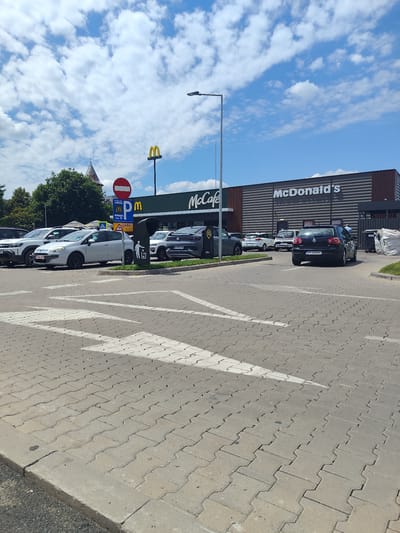 The image shows the exterior of a McDonald's restaurant in Arad, Romania, on a sunny day with a partially cloudy sky. The McDonald's building is modern, with a dark facade and the iconic golden arches visible on the roofline. The restaurant also features a McCafe section. In front of the restaurant is a parking lot paved with interlocking concrete blocks. Several cars are parked in the lot, including a black Volkswagen Golf, a silver Fiat Punto, and a white SUV. A pedestrian crossing with white arrows is painted on the pavement. In the foreground, the interlocking concrete blocks and a curb are visible. Traffic signs, including a red circular "no entry" sign and a blue parking sign with a "P" symbol, are present near the entrance of the parking lot. Green trees and a building with a spire, possibly a church, are visible in the background. The scene appears to be a typical commercial area in the city of Arad.