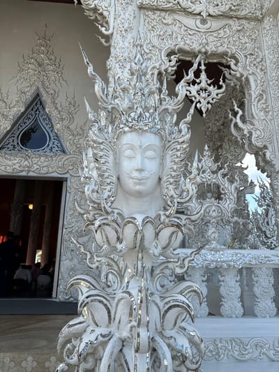 The scene displays a detailed exterior view of Wat Rong Khun, commonly known as the White Temple, located in Ban Rong Khun, Thailand. In the foreground, a large, intricately sculpted white bust of a deity or mythical figure is centrally positioned. This figure features a serene face with closed eyes, a delicate mustache, an elaborate multi-layered crown, and extensive flame-like embellishments extending from the head and neck. The entire sculpture, including its lotus-style base, is composed of white material extensively inlaid with small, reflective mirror pieces. To the left, an open temple entrance reveals a dimly lit interior with dark vertical columns and several indistinct human figures seated or kneeling on the floor. Above this entrance, a triangular window with a dark, reflective surface is visible, framed by ornate white stucco work. The temple's facade, extending to both the left and right of the bust, consists of complex, swirling white architectural elements also embedded with numerous mirror fragments. A glimpse of blue sky is visible through an opening in the ornate structure to the far right, indicating an outdoor, daylight setting.