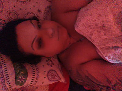 The image displays a close-up, sideways view of a person's face and upper body. The person has dark hair and appears to be lying down. They are wearing a light-colored, patterned garment draped over their shoulder and chest. The background is a reddish-pink textured surface with subtle, darker patterns resembling paisley or floral motifs. The lighting is dim and casts a warm glow over the scene. The image is grainy and has a low resolution, making fine details indistinct. There is no discernible information or context that places the image in the city of Komotini, Greece.