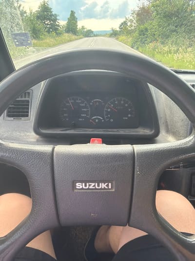 The image, rotated 90 degrees clockwise, captures the interior of a car, focusing on the steering wheel and dashboard, with an expansive view of the road and landscape outside. A person's bare legs are visible behind the steering wheel, indicating a driver or passenger wearing shorts or a skirt.

The prominent black, textured steering wheel features a "SUZUKI" logo at its center. Behind it, the instrument cluster displays a speedometer showing 0 KM/H, a tachometer indicating approximately 1000 RPM, a fuel gauge that is about half full, and a temperature gauge reading low. A red hazard button is visible to the right of the gauges, and a wiper control stalk with settings "MIST", "LO", and "HI" is positioned higher up. An air vent is located on the lower left of the dashboard.

Through the windshield, a sunny daytime scene unfolds. The car appears to be on an asphalt road, flanked by abundant green trees and vegetation. Distant hills or mountains rise under a sky with scattered clouds, suggesting a rural or mountainous setting. A small, partially illegible sticker is affixed to the lower left portion of the windshield. The scene depicts a drive through Stanyantsi, Bulgaria.