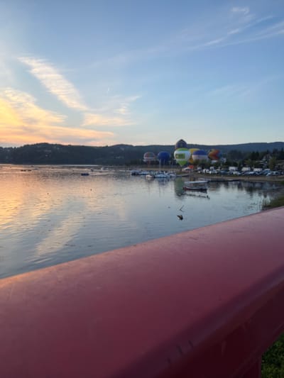 The image shows a wide body of water, likely Lake Rożnowskie in Gródek nad Dunajcem, Poland, at sunset. The sky is painted with soft orange and yellow hues near the horizon, transitioning to a clear blue with wispy clouds. Several hot air balloons, some in vibrant colors and patterns, are visible in the distance, hovering above the treeline that borders the lake. The water reflects the colors of the sky and the surrounding landscape, creating a serene atmosphere. Small boats are scattered across the water, and a marina with docked vessels is visible along the shore. In the foreground, a red railing is partially visible, suggesting the photo was taken from a bridge or a similar elevated vantage point. The overall scene depicts a peaceful evening at a popular lakeside destination.