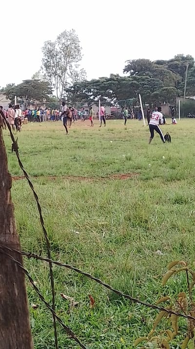 The image shows an outdoor volleyball game underway in the Siaya Township ward, Kenya, viewed from behind a partial barbed wire fence. In the immediate foreground, a wooden fence post is visible on the left, with several strands of barbed wire extending horizontally across the lower left and central sections of the frame. The ground is covered in green grass, interspersed with patches of exposed dirt and some scattered debris.
In the midground, a volleyball net, appearing informal and supported by two white posts, spans the grassy playing area. Approximately ten individuals are actively engaged in the game. One player, prominently positioned in the right midground, wears a white shirt with the number "23" on the back and black athletic pants, captured in a dynamic pose. A dark object, possibly a bag, rests on the ground near this player. Other participants are visible near the net, dressed in various casual attire including pink and green shirts.
In the background, a crowd of approximately 20-30 spectators is gathered behind a discernible boundary line, observing the game. Beyond the spectators and game area, a dense line of green trees forms the horizon. Scattered amongst the trees or behind them are indistinct structures. The sky is uniformly light, indicating an overcast daytime condition.