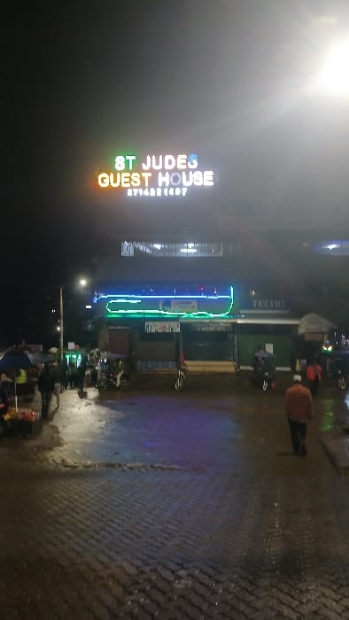 This image displays a nighttime street scene in Kisii Central ward, Kenya. The foreground consists of dark grey interlocking pavers, wet and reflective from recent moisture. In the mid-ground, a multi-story building dominates, featuring a prominent, brightly illuminated sign on its upper facade that reads "ST JUDES GUEST HOUSE" in multi-colored letters (green, orange, blue, white), with the contact number "0714301887" displayed below. Below this sign, a segment of the building exhibits horizontal blue and green neon strip lighting. A "TECNO" logo is visible on the building's facade to the right of the neon lights. At street level, on the far left, a person stands under a blue umbrella next to a street stall with items displayed in red containers. Several individuals are present on the wet pavement, including a figure wearing a light-colored cap and a brown-orange jacket, walking away from the camera towards the right side of the frame. Multiple motorcycles are visible, some stationary and others with riders. A bright streetlight on a pole emits a strong glow from the left side of the scene.