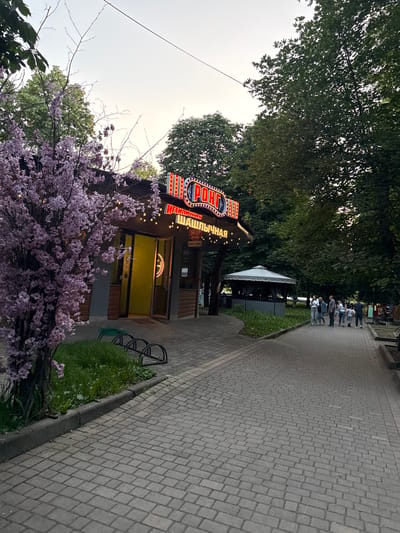 An outdoor scene at dusk features a walkway paved with grey bricks leading away from a small building with a neon sign. The building appears to be a food establishment, with a bright yellow interior visible through its entrance. The neon sign, illuminated with red and blue lights, displays text in Russian, reading "РОНД" and below it, "НЕТИПИЧНАЯ ШАШЛЫЧНАЯ". A decorative tree with pink blossoms stands to the left of the building. Several people, appearing to be a group of tourists, are walking along the brick pathway. In the background, trees and a covered outdoor seating area are visible. The sky is a pale grey, suggesting the transition from day to night.