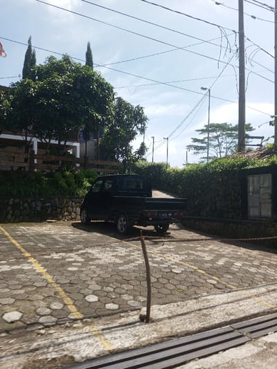 The image displays an outdoor scene in Beteng_Pangkhar_Somthrang, Bhutan, under a clear, bright sky. A black small-sized pickup truck, bearing an Indonesian license plate 'F 9241 MNN' on its rear, is parked on a paved surface made of grey and off-white hexagonal interlocking concrete pavers. The truck faces away from the viewer, angled slightly to the right, with a small quantity of light-colored material, possibly sand or gravel, present in its open cargo bed. Yellow painted lines mark a parking space on the left side of the pavement. A dark-colored metal chain extends horizontally across the lower middle ground, connected to a partially visible vertical post. In the immediate foreground, a thin, dark stick-like object protrudes vertically from the pavement, and a metal drainage grate is visible at the bottom right.
On the left side of the frame, a multi-story building is partially visible, featuring white railings on a balcony or porch above a light-colored wooden fence. Dense green foliage from several trees, including tall, slender coniferous trees and broader, round-canopied trees, surrounds this building. To the right of the parked truck, against a dark stone retaining wall covered in green shrubs, stands a dark rectangular structure or shed with a silver or metallic double door. Multiple dark utility poles rise in the mid-ground and background, supporting numerous overhead power lines or cables that crisscross the upper sky. Several streetlights are affixed to these poles. In the background, further trees with distinct horizontal branching patterns are visible against the sky. No individuals are present in the scene, and no specific actions or interactions are depicted.