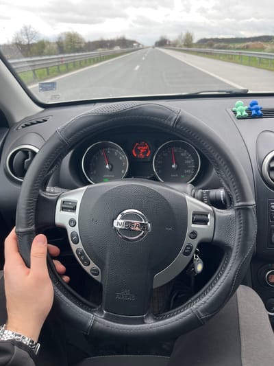 A view from inside a Nissan vehicle, the steering wheel prominently displayed with the Nissan logo at its center. The driver's hand is visible gripping the wheel, and a silver wristwatch adorns their wrist. The dashboard includes a speedometer reading approximately 80 km/h, alongside other vehicle controls. Outside the car, a highway stretches into the distance under an overcast sky. Given the data embedded in the image, the mileage of the car is 104589 km, the temperature is 38.4°C, and the time is 1:13. The location is purportedly Trilistnik, Bulgaria.