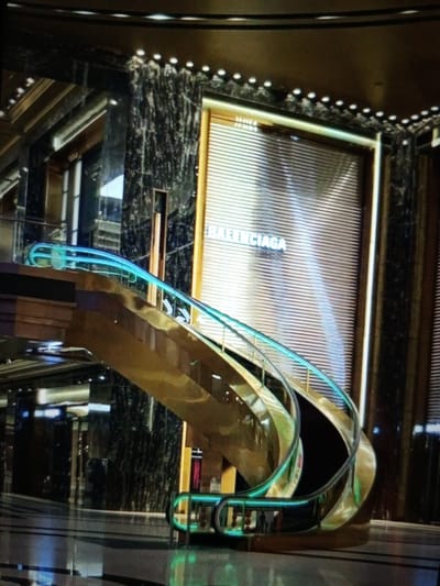 The image shows a luxurious, modern interior with a prominent spiral escalator. The escalator has a golden railing and is illuminated with turquoise LED lights. The surrounding architecture features dark, polished marble columns and a large, ribbed facade with the "BALENCIAGA" logo visible. The flooring appears to be dark and reflective. The overall impression is one of an upscale retail or commercial space. The image does not provide any specific information about the city of Mutum Mbiyu, Nigeria, or any people in the scene.