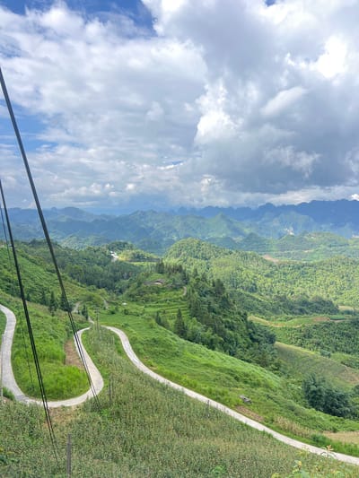 The image depicts a mountainous landscape with lush green vegetation and a winding road. In the foreground, a paved road curves sharply down a grassy slope. Two thick, black cables run vertically along the left side of the image, partially obscuring the view. The road is surrounded by dense green foliage, including tall grasses and some trees.
Further in the background, the landscape transitions into rolling hills covered in a variety of green vegetation, including what appears to be terraced fields. The hills are densely forested, with numerous trees creating a textured green carpet. In the distance, a range of hazy, blue-hued mountains stretches across the horizon, their peaks fading into the atmospheric perspective. The sky above is partly cloudy, with patches of blue sky visible between dramatic, greyish-white clouds that cast shadows on the landscape.
The scene is clearly a rural, mountainous region. While the prompt specifies Quản Bạ Commune, Vietnam, the image itself does not contain any explicit markers to confirm this location. However, the topography and vegetation are consistent with mountainous areas in Southeast Asia. There are no people or animals visible in the image. The overall impression is one of natural beauty and dramatic scenery.