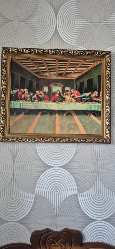 A framed painting depicting "The Last Supper" hangs on a wall with a decorative, textured wallpaper. The painting shows Jesus and the twelve apostles seated at a long table. The wallpaper features a pattern of concentric arcs in varying shades of gray. The bottom portion of the image shows a carved wooden headboard. The location provided, Metsamor, Armenia, is not depicted in the image.