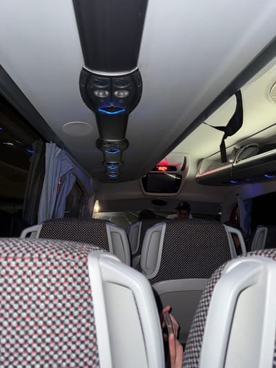 The image depicts the interior of a passenger bus or coach, observed from a forward-facing perspective within the passenger cabin. The location is Shoul السهول, Morocco. The scene is illuminated by artificial lighting, primarily from a central ceiling fixture strip featuring multiple circular units emitting blue ambient light, in addition to general cabin lighting.

The bus features rows of passenger seats upholstered in a distinctive black, white, and red checkered fabric, with light gray or white plastic handles on the seatbacks. Overhead luggage compartments are visible on both sides, with a black fabric strap hanging from the compartment on the right. In the forward section of the cabin, a digital display shows "0:04" in red numerals, positioned above a small screen. Window curtains, light gray in color, are visible pulled back on the left side.

At least three individuals are present. In the immediate foreground, a pair of hands belonging to an unseen passenger is visible, holding a light-colored smartphone. Further into the cabin, behind multiple rows of seats, the upper body of one individual wearing a dark cap is discernible, looking towards the front of the bus. Another person's head is partially visible to the right of the person in the cap. The exterior environment, visible through the front windshield, is dark, showing faint lights consistent with a road during nighttime or very early morning travel.