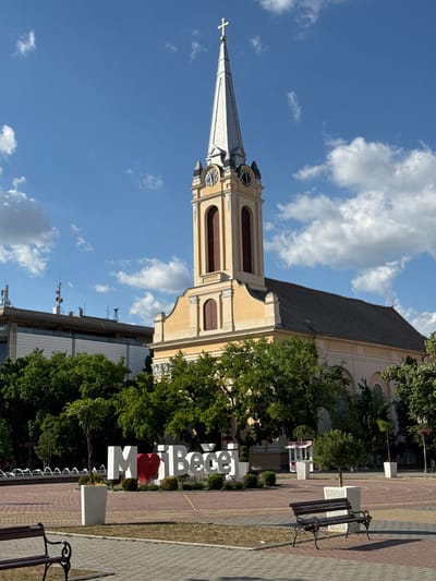 This image captures a wide shot of a town square dominated by a large, light yellow church under a bright blue sky with scattered white clouds. The church features a tall, silver-colored spire topped with a golden cross. Below the spire, the church tower has two visible clock faces, each displaying approximately 11:55. Rectangular, arched windows with reddish louvers are visible on the tower. The main body of the church has a dark grey roof and several arched windows, some partially obscured by trees.

To the left of the church, a modern building with a flat roof is visible. This building has multiple antennas and communication equipment mounted on its roof. The facade below the roof appears to have white, horizontal paneling.

In the foreground and midground, a paved town square is visible, patterned with different shades of brown and reddish pavers. Several park benches with dark metal frames and wooden slats are placed on the paved areas. White, rectangular planters containing small trees and shrubs are scattered across the square. A large, white "I ❤️ Bečej" sign is prominently displayed in the center-left of the square, with a red heart substituting the word "love." In the far-left background, a series of fountains creating water arcs can be partially seen. Lush green trees are abundant around the perimeter of the square and in front of the church. The scene is brightly lit by sunlight, casting distinct shadows from the benches and planters. No people are visible in the image.

The location is the town square of Bečej Municipality, Serbia, as indicated by the "I ❤️ Bečej" sign.