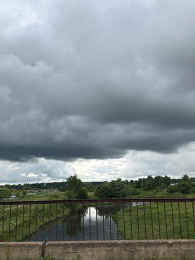 This media file depicts a scene from Timofeevo, Russia, with a dominant stormy sky. The composition is vertically oriented, with a dark, ominous cloud bank occupying the left half of the frame. This cloud bank appears dense and foreboding, suggesting an impending storm or the aftermath of one.

To the right of the storm clouds, the sky transitions to a brighter, more scattered cloud formation, with hints of sunlight breaking through. Below this sky is a landscape view. A rusty metal railing runs along the right side of the image, in front of a concrete embankment. Beyond the railing, a narrow body of water, likely a river or canal, flows. Its surface reflects the sky and the surrounding greenery.

The banks of the water are lined with lush green grass and trees, forming a dense, natural border. The trees appear to be deciduous, and their foliage is thick, indicative of a summer or late spring season. In the distance, beyond the immediate treeline, some buildings or structures are visible, hinting at a settlement or rural area.

The overall atmosphere is one of dramatic weather. The contrast between the dark storm clouds and the brighter, partly cloudy sky creates a sense of tension. The natural elements – water, grass, trees – are rendered in subdued tones, consistent with overcast or transitioning weather conditions. There is no visible text in the image. The time of day is not explicitly stated but the quality of light suggests it could be late afternoon or early evening, when storm clouds often gather.