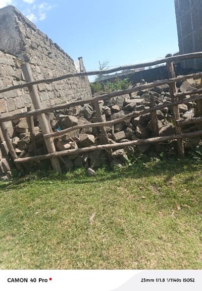 The image displays an outdoor daytime scene located in Nakuru East ward, Kenya. The foreground consists of green grass with visible patches of drier vegetation. A rustic, horizontally slatted wooden fence, constructed from rough-hewn posts and rails, runs across the mid-ground, angled slightly from the lower left to the upper right. Immediately behind this fence, a large, disordered pile of dark grey, irregularly shaped rocks and broken concrete debris is present, forming a substantial barrier. To the left, a wall constructed from large, rough-faced grey stone or concrete blocks rises, indicating a building structure. In the background, under a clear blue sky with scattered white clouds, other elements are visible: a corrugated metal chimney extending from a roofline on the middle-left, and a taller structure made of light-colored concrete blocks on the far right. A utility line or wire stretches horizontally across the distant background, and a faint outline of a distant object is discernible on the horizon. No individuals, animals, or explicit actions are depicted. The scene presents an unoccupied, semi-developed or transitional outdoor area.