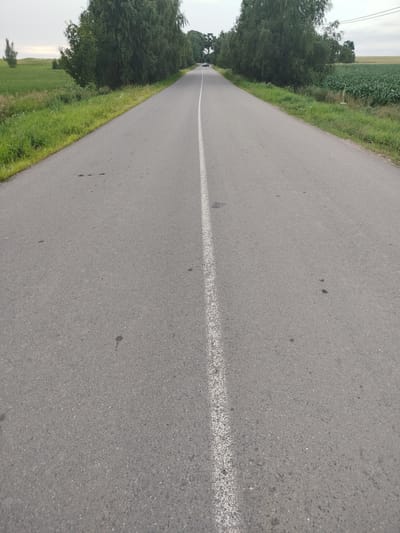 A two-lane asphalt road stretches into the distance, flanked by lush green grass and trees on either side. In the background, a white car is visible traveling away from the camera. The sky is overcast, suggesting a cloudy day. The road appears to be a rural route, possibly located in the vicinity of Крупіцкі сельскі Савет, Belarus, given the agricultural fields visible. The road surface shows signs of wear, with some small dark patches and faint marks scattered across its width. The white line in the center of the road is faded in some areas.