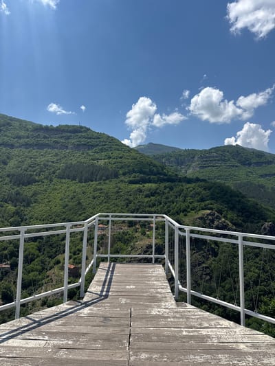 This image captures a panoramic view from a wooden viewing platform located in Zasele, Bulgaria. The platform, constructed from weathered light brown wooden planks with visibly peeling paint, extends into the foreground. A white metal railing, featuring horizontal bars and vertical suspension cables, delineates the edge of the platform and the viewpoint. Long, distinct shadows of the railing are cast across the wooden surface, indicating bright sunlight from the upper left.
Beyond the railing, a vast, deeply forested mountainous landscape unfolds under a clear blue sky dotted with fluffy white cumulus clouds. The mountains are densely covered in vibrant green foliage, with varying shades indicating different types of trees and terrain features. A notable, prominent flat rock face is visible on the side of one of the mountains. On the peak of a distant mountain, a slender telecommunications tower or mast is discernible. Scattered among the trees in the mid-ground and far distance, a few small houses or buildings with reddish roofs can be seen, suggesting human habitation within the valley. No people are present in the scene.