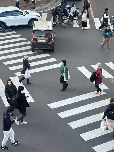 This image presents an overhead view of a street intersection in Tokorozawa, Japan, identified by the "所沢" marking on the license plate of a brown compact car (plate number 580 さ 11-22). The scene captures a busy moment with multiple pedestrians utilizing a marked crosswalk and several vehicles.

In the upper left, a white SUV is partially visible. A brown compact car is positioned on the crosswalk, displaying a green and yellow "wakaba mark" (beginner driver symbol) on its rear window. Further up and to the right, several individuals are cycling on bicycles, some equipped with child seats.

Approximately 15 individuals are depicted, all appearing to be pedestrians actively crossing the intersection. Their attire is consistent with cool weather, featuring coats, sweaters, and long pants. Many individuals are wearing white disposable face masks. Notable pedestrians include:
*   A woman in a dark coat, light-colored beanie, and dark boots, carrying a light-colored bag, walking left across the crosswalk.
*   A woman in a green sweater, dark pants, and dark boots, carrying a white tote bag, walking right across the crosswalk.
*   An older woman with grey hair, wearing a red tunic and dark pants, carrying a dark garment, walking left across the crosswalk.
*   A man in a teal jacket and tan pants, holding a mobile phone, walking right.
*   Several individuals, including a man with a black backpack and a man in a black jacket and white pants, are observed looking at or holding mobile phones while walking.

The environment consists of an asphalt road with clearly visible white striped crosswalks. The lighting is natural daylight, suggesting an overcast sky or an early/late hour of the day due to the soft shadows.