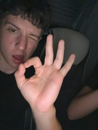 A young person is in a car, possibly at night. The person is making a circle shape with their thumb and forefinger, winking at the camera. The interior of the car is visible, including the seat headrest. The location is not Vinitsa, Bulgaria.