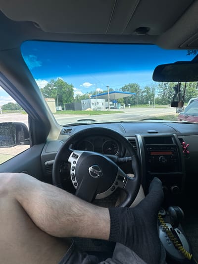 The image shows the interior of a car, viewed from the perspective of the driver. The driver's right foot, wearing a black sock, is resting on or near the steering wheel. The steering wheel has the Nissan logo on it. The dashboard contains a radio and ventilation outlets. Outside the car, through the windshield, a bright blue sky with scattered white clouds is visible, suggesting it is daytime. Trees and buildings are also visible, including what appears to be a gas station with a blue canopy. The overall scene is a snapshot of someone inside a vehicle, possibly parked or at a standstill. The lighting and clear sky indicate fair weather.