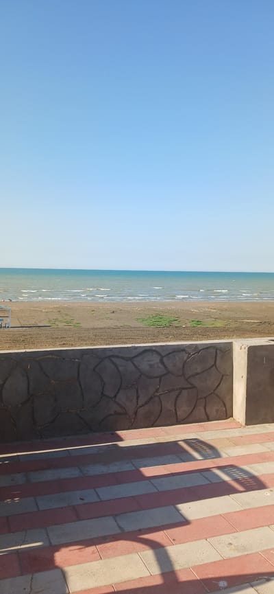 This is a landscape image of a beach on a clear, sunny day. The scene is divided into three main parts: a vast expanse of clear blue sky on the left, the ocean and sandy beach in the middle, and a paved walkway with patterned tiles on the right.
The sky is a consistent, light blue, suggesting it is daytime with no clouds. The ocean is a calm, greenish-blue with small waves gently lapping at the shore. The beach is a light brown sand, with some patches of green grass growing near the edge of the water. There are a few small figures of people visible on the sand and in the water.
The walkway on the right is constructed with alternating red and light-colored rectangular tiles, laid in a striped pattern. A section of the walkway features a different paving pattern with dark, irregularly shaped stones arranged to resemble a mosaic. The shadows cast by an unseen object, possibly a railing or fence, fall across the tiled area, indicating a low angle of the sun, suggesting either early morning or late afternoon.
The location is identified as Sumqayit, Azerbaijan. The overall mood is peaceful and serene, typical of a beach setting on a pleasant day. No text is visible in the image.