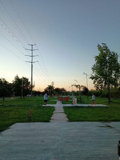 This media file depicts an outdoor park or green space in General Pacheco, Argentina, during what appears to be late afternoon or early evening, possibly near sunset. The sky, a clear expanse of light blue overhead, graduates to a softer, yellowish hue closer to the horizon.
A wide, paved pathway, likely concrete or asphalt, stretches through the center of the frame, bordered by well-maintained green grass. Along the pathway, several park amenities are visible, including benches and red structures that could be exercise equipment or playground elements, leading into the distance. A bicycle is parked near one of the benches. Trees with varied green foliage line the background and dot the landscape, contributing to the park setting.
Prominently featured on the left side of the image is a large lattice-style metal power pole, from which numerous power lines extend horizontally across the sky. Other, simpler utility poles are also visible further along the path. Streetlights are intermittently placed along the pathway.
Two individuals are present: one person, dressed in a light-colored top and dark shorts, stands on the grassy area near the foreground path. Another person, also in light clothing, is seen further down the path near the red structures. Their activity appears casual, suggesting leisurely use of the park. The overall scene conveys a calm and peaceful atmosphere, with no specific event taking place. There is no visible text in the image.
