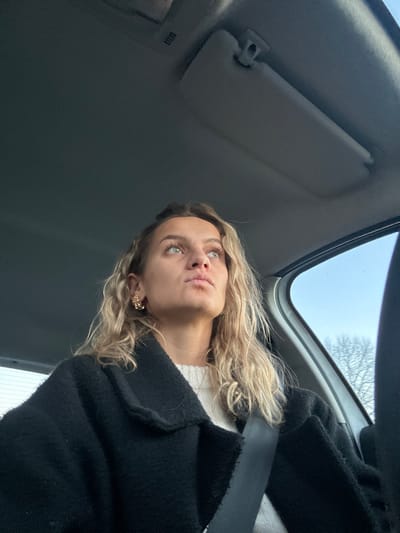 A low-angle shot from inside a car shows a woman with blonde, curly hair. She wears a white top and a black coat, secured with a seatbelt. Her face is turned upwards, lips slightly pursed, and her eyes are focused out of frame. In the background, a portion of the car's interior is visible, including the sun visor, roof, and a sunroof offering a glimpse of the sky and tree branches.