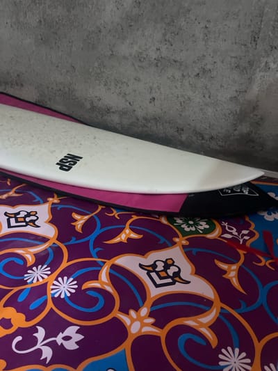 In Desa Batu Layar, Indonesia, two surfboards are displayed against a textured grey wall. The prominent upper surfboard is white, featuring the black "NSP" logo and a visible wax pattern on its surface. It rests upon a second surfboard or board bag that is predominantly pink with a black section visible on the right, bearing partial text "LIVE" and "TOP." Both surfboards are positioned on a vibrant, multi-colored mat or rug, which displays an intricate pattern of purple, orange, blue, and white, with floral and scroll motifs. No individuals are present in the scene.