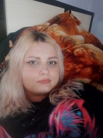A woman with blonde hair and light skin is lying on a bed. She is wearing a black shirt with a colorful feather-like design. The background is a patterned comforter and a wall. The image appears to be taken indoors. The location of Изыхские Копи, Russia is irrelevant to the visual information presented in the image.