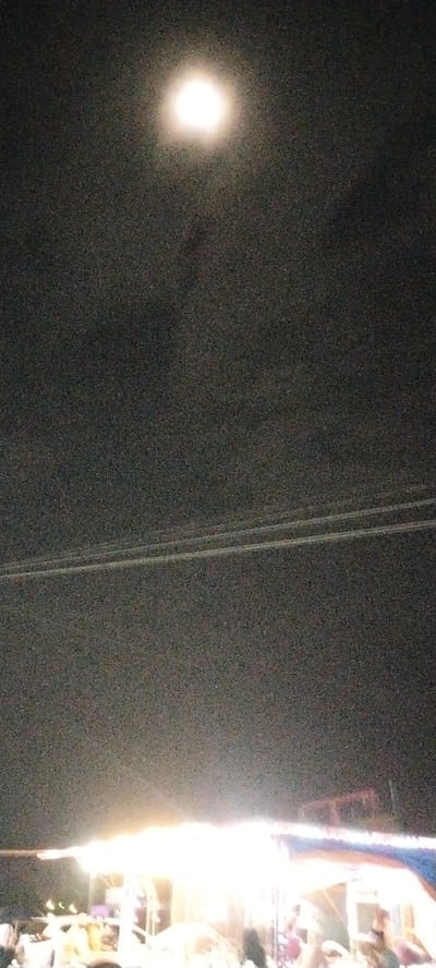 The image depicts a nighttime scene under a full moon. The sky is dark and grainy, with the bright, blown-out moon positioned in the upper center. Below the moon, two thin, parallel lines are visible, likely power lines or wires, stretching horizontally across the frame.
The lower portion of the image is illuminated by bright, artificial lights emanating from what appears to be a stage or a series of stalls, possibly at an event or festival. Silhouettes and blurred figures of people are present at the bottom, suggesting a gathering or crowd. The lighting is harsh and overexposed in this area, washing out details of individuals and structures. The overall impression is of an outdoor event taking place at night, with the full moon and illuminated structures being the primary visual elements. The specific location in Gampongpaya, Indonesia, is not visually identifiable within the image itself.