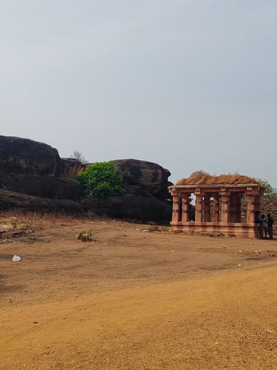 The image depicts a small, open, red sandstone pavilion-like structure in Vamasamudram, India. The structure has a straw-covered roof and is located on a reddish-brown, dusty, and barren landscape. To the left are large, dark rocks with a small patch of green foliage. Two individuals are standing near the right side of the structure. A small patch of green foliage sits on the barren ground in front of the rocks. The sky is a pale greyish-blue. The overall impression is one of a dry and sparsely vegetated environment.