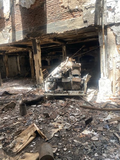 The image depicts a building interior devastated by fire. Burned rubble and debris cover the floor. Walls are charred, and some areas show exposed brickwork. The structure appears unstable, with damaged beams and supports visible. A section of what may have been a staircase or multi-level structure is partially collapsed and severely burned. The overall impression is one of extreme destruction.