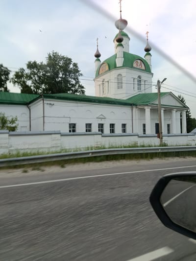 The image shows a white building with a green roof, topped with several domes and crosses, in Khoteichi, Russia. The building appears to be a church. It is flanked by trees and surrounded by a white fence. A road with a metal guardrail runs in front of the building, and a car's side mirror is visible in the foreground. The sky is overcast, and a few birds are flying. The scene is captured from a moving vehicle, indicated by the blur in the foreground and the reflection in the side mirror.