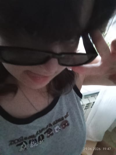 A person, wearing dark sunglasses and a grey tank top with a black trim, is captured in a close-up, slightly blurry selfie. The tank top has a printed design of cartoon dogs and some text. The person's face is partially obscured by the sunglasses and a dark object, possibly a hat or hair accessory, is on their head. Their right hand is raised, with fingers extended, as if posing or waving.
The background appears to be an indoor setting with a window and a radiator visible. There's a white curtain or fabric to the right of the person. The lighting suggests it might be daytime, possibly late afternoon as indicated by the visible timestamp. The overall mood of the image is casual and lighthearted, with the gesture and slight smile suggesting a playful moment.
The visible text on the tank top includes "2002" and some stylized images of dogs. A timestamp in the bottom right corner reads "29.06.2026 19:47". The location context provided is Tyrnyauz, Russia.