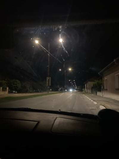 The image, taken at night, depicts a street scene from the interior of a vehicle, likely in the city of Yasen, Bulgaria. The perspective is from the driver's seat, looking forward through the windshield, which shows some streaks and smudges.
The road ahead is illuminated by streetlights, creating bright points of light and reflections on the wet asphalt. Several streetlights are visible along the left side of the road, casting a yellow glow. On the right side of the road, houses with illuminated windows are visible. The houses appear to be residential, with one closer to the foreground showing a pale facade. There is a sidewalk along the right side of the road, bordered by white curb stones.
Vegetation, including trees and bushes, is present on the left side of the road, behind a low fence or wall. Parked cars are dimly visible on the left side of the street. In the distance, more streetlights line the road, creating a sense of depth. Headlights of an oncoming vehicle are visible further down the street. The overall atmosphere is that of a quiet, suburban or semi-urban street at night. The dashboard of the car is visible in the foreground, mostly in shadow.