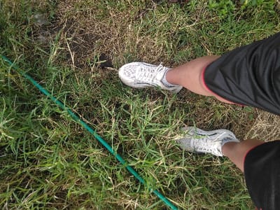 This overhead shot depicts the lower legs and feet of an individual standing on uneven ground. The person wears black athletic shorts with red trim, white socks, and white athletic shoes with grey accents, identifiable as Asics. The ground consists of a mix of green and dry brown grass, with visible patches of dark soil. A green hose traverses diagonally across the lower left section of the frame, partially concealed by vegetation. No specific landmarks or architectural features are present to determine the precise location within Røros, Norway.
