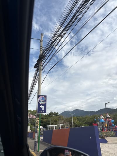 The image shows a view from inside a car in San Rafael, Costa Rica (latitude: 9.938, longitude: -84.153). A pole with a blue and white sign with the text "Baga H. Usa" and an arrow pointing left is attached to a pole. The sign indicates that it is 170 meters to the destination. There are a lot of power lines attached to the pole. In the distance, there is a blue and orange concrete barrier and a playground behind it. The area is covered with green vegetation and there is a mountain range in the background. The sky is partly cloudy.