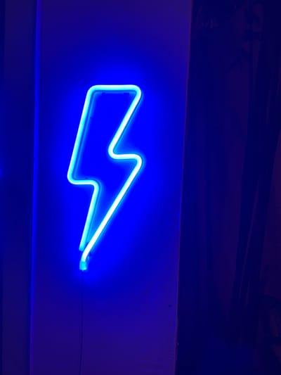 A brightly illuminated blue neon lightning bolt sign is mounted horizontally on a textured, white surface, likely a wall. The surrounding environment is primarily dark blue, with a darker area above the sign suggesting a ceiling or overhang. A thin wire extends from the right end of the sign, indicating its power source. The overall impression is one of artificial lighting in a possibly confined space. Location in Buduk, Indonesia is unknown.