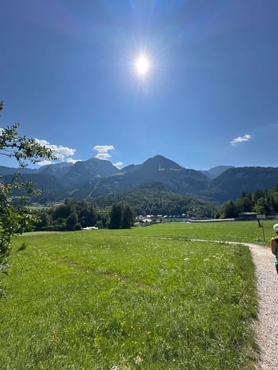 This is a wide-angle, eye-level, outdoor photograph taken on a sunny day in Schönau am Königssee, Germany. The scene depicts a vibrant green meadow dotted with small white and yellow wildflowers, leading to a light-colored gravel path on the right. In the background, lush green trees cover rolling hills, which transition into majestic, imposing mountains with visible ski slopes. A small village with buildings and a body of turquoise water are nestled at the base of the mountains. The sky is a clear, bright blue, dominated by the sun, which shines intensely and creates a lens flare effect. A person wearing a white hat and a teal top is visible on the right side of the path, walking away from the camera. The image captures a scenic view of the Bavarian Alps, a popular tourist destination known for its natural beauty and outdoor activities.