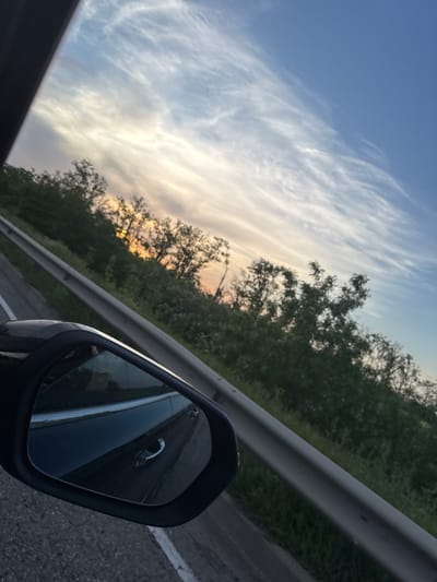 The image shows a view from a car, likely on a highway, in or near Fastovetskaya, Russia. The view is angled, and includes the car's side mirror, a guardrail, trees, and the sky. The side mirror reflects the side of the car and the road behind it. The guardrail runs horizontally across the image. Trees line the other side of the guardrail. The sky is visible with wispy clouds, suggesting either dawn or dusk. The road surface is visible in the lower right of the image.