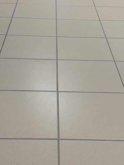The image shows a close-up, angled view of a beige tiled floor with dark gray grout lines. The tiles are square and laid in a grid pattern. The lighting suggests an indoor environment with some overhead light source creating subtle reflections on the smooth tile surfaces. There are no people, objects, or actions depicted in the image. The location is identified as Malmö-Sturup, Sweden, but this specific context is not visually evident from the image itself.