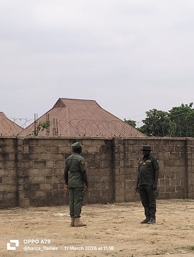 The image displays an outdoor scene in Gidan Kado, Nigeria, featuring two uniformed males standing on dry, light-brown earth.
In the foreground, the ground is sandy and sparsely covered. In the midground, two adult males, both wearing matching short-sleeved green uniforms and caps, are present. The man on the left stands with his back to the viewer, wearing light brown boots. The man on the right faces the viewer, his head slightly turned towards the left man, wearing dark shoes and a dark cap, with an insignia visible on his left sleeve.
Behind the individuals, a weathered concrete block wall, approximately two meters in height, stretches across the frame. Multiple coils of barbed or razor wire are affixed to the top of this wall, running its length.
In the background, behind the security wall, a reddish-brown corrugated metal roof of a building is partially visible on the left. To the right, green tree foliage extends above the wall. The sky overhead is overcast and light grey. Both men are standing still, not engaged in active movement or direct interaction.