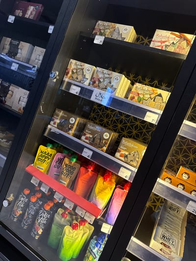 The image shows a refrigerated display case filled with food products, likely in a supermarket in Ilidža, Bosnia and Herzegovina. The products include various types of packaged sweets, notably "Rahatlokum" (Turkish delight), and pouches of nutritional drinks or mixes. Some of the products have Arabic script on their packaging, indicating an international selection. The display case is well-lit, and the glass front allows for a clear view of the merchandise. The overall impression is one of a clean, organized retail environment offering a diverse range of food items. There are no people visible in the image.